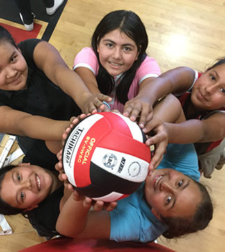 Female students hold up a volleyball in a gym