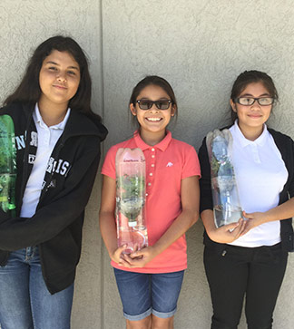 Three female students pose in front of a wall as they hold water bottle gardening projects